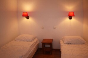 A room with two single beds, each with a white pillow and a red wall lamp. There is a small wooden nightstand between the beds.