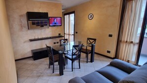 A modern living room with a glass dining table, dark chairs, a sofa, a TV mounted on the wall, and a window with curtains. - Two-room apartament in heart of tuscany (Montopoli)