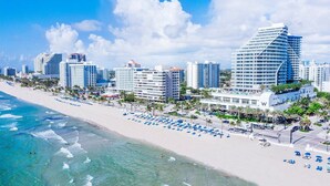 A beachfront hotel with multiple high-rise buildings, a sandy beach, and numerous blue umbrellas.