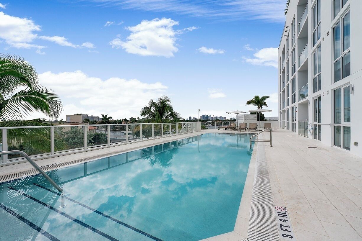 A rooftop swimming pool with clear blue water, surrounded by a white building with large windows and a palm tree.