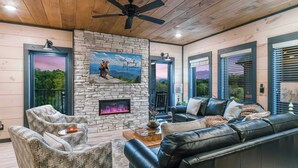 A cozy living room with a stone fireplace, leather sofa, and a ceiling fan. - 22 Guests | Luxury Chalet | Indoor Pool | Mtn View (Sevierville)
