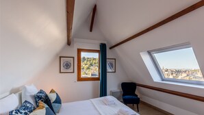 Un dormitorio con cama, una silla, un ventanal con vistas y dos cuadros en la pared.