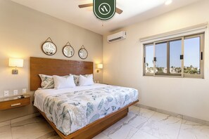 A bedroom with a large bed, a wooden headboard, wall-mounted lamps, and a window with a view. - Maia Cabo by Naya Homes (Cabo San Lucas)
