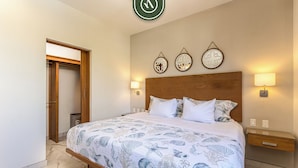 A bedroom with a bed, a wooden headboard, wall-mounted lamps, and a door leading to another room. - Maia Cabo by Naya Homes (Cabo San Lucas)