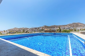 A swimming pool with clear blue tiles, surrounded by a concrete deck, set against a backdrop of a mountainous landscape and residential buildings. - Maia Cabo by Naya Homes (Cabo San Lucas)