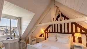 Een slaapkamer met een schuine kap, een bed met witte lakens, bedlampen, een nachtkastje en een raam met uitzicht op een stad.