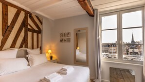 A hotel room with a large bed, a wooden ceiling, a window with a view of buildings, and a mirror on the wall. - Dolphin view - 1st floor - Charming, comfortable flat on the port (Honfleur)