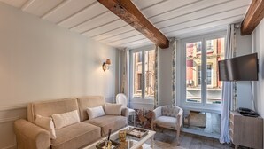 A living room with a beige sofa, a white armchair, a glass coffee table, and a wooden floor. There is a large window with a view of buildings outside. - Dolphin view - 1st floor - Charming, comfortable flat on the port (Honfleur)