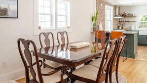 A dining area with a wooden table and chairs, a kitchen in the background, and a cityscape view through the window. - Downtown Greenway Getaway (Raleigh)