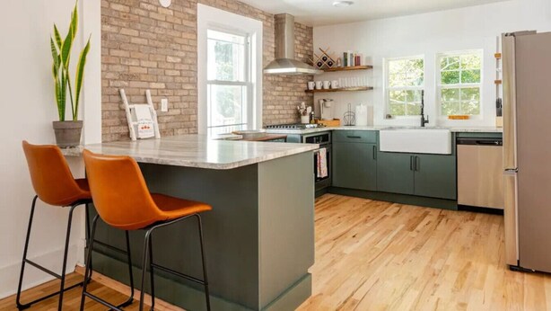 A modern kitchen with a brick accent wall, a central island with bar stools, and a view of the outdoors through large windows. - Downtown Greenway Getaway (Raleigh)