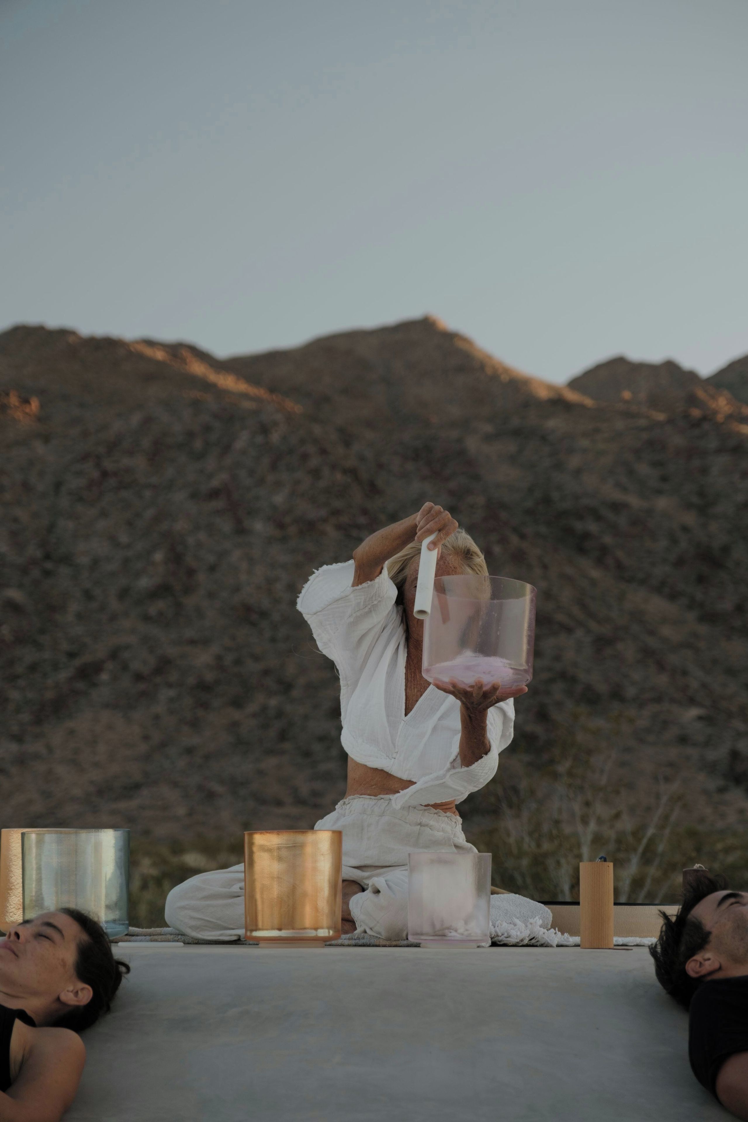 A person in a white outfit performing a yoga pose with a clear glass in hand, seated on a concrete surface with a mountainous backdrop.