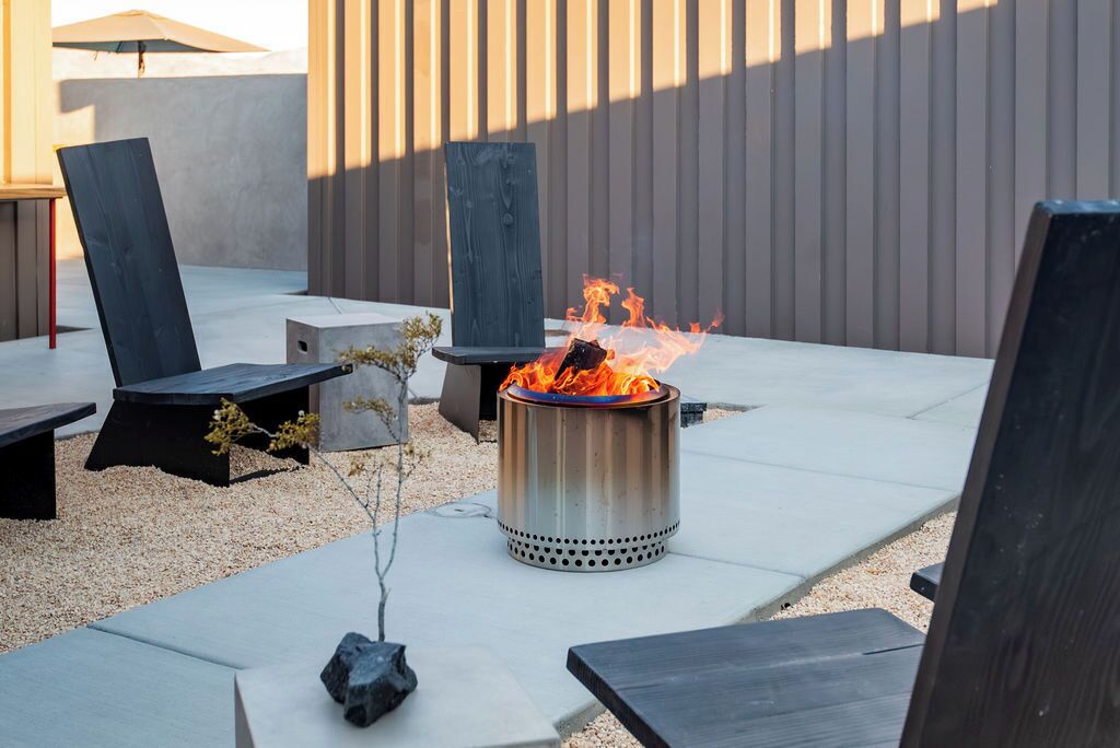 Un patio con un pozo de fuego de metal, sillas de lounge negras y una mesa de hormigón.
