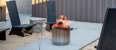 Un patio con un pozo de fuego de metal, sillas de lounge negras y una mesa de hormigón.