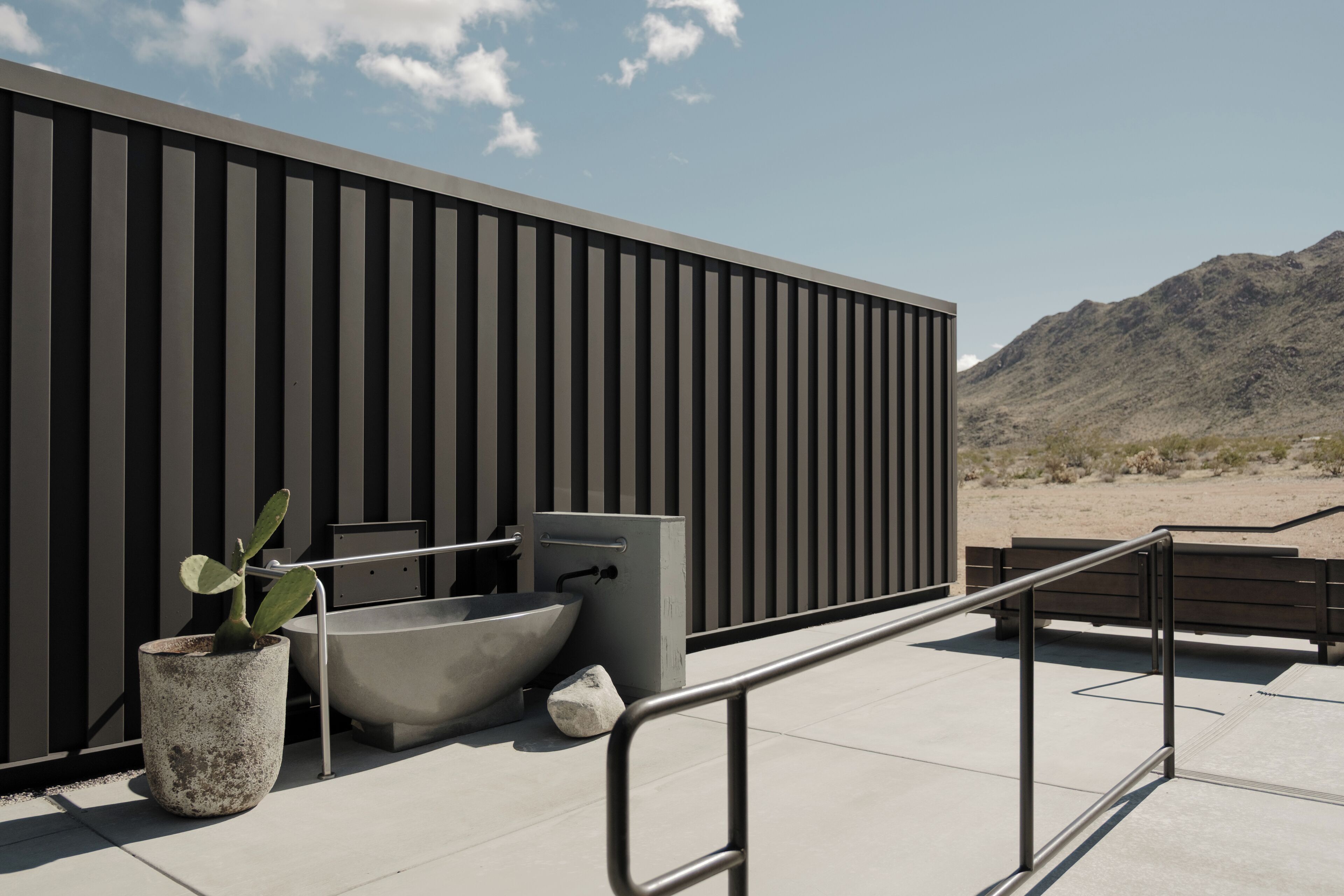 A modern outdoor bathroom with a large planter, a bathtub, and a sink against a vertical panel wall, overlooking a desert landscape.