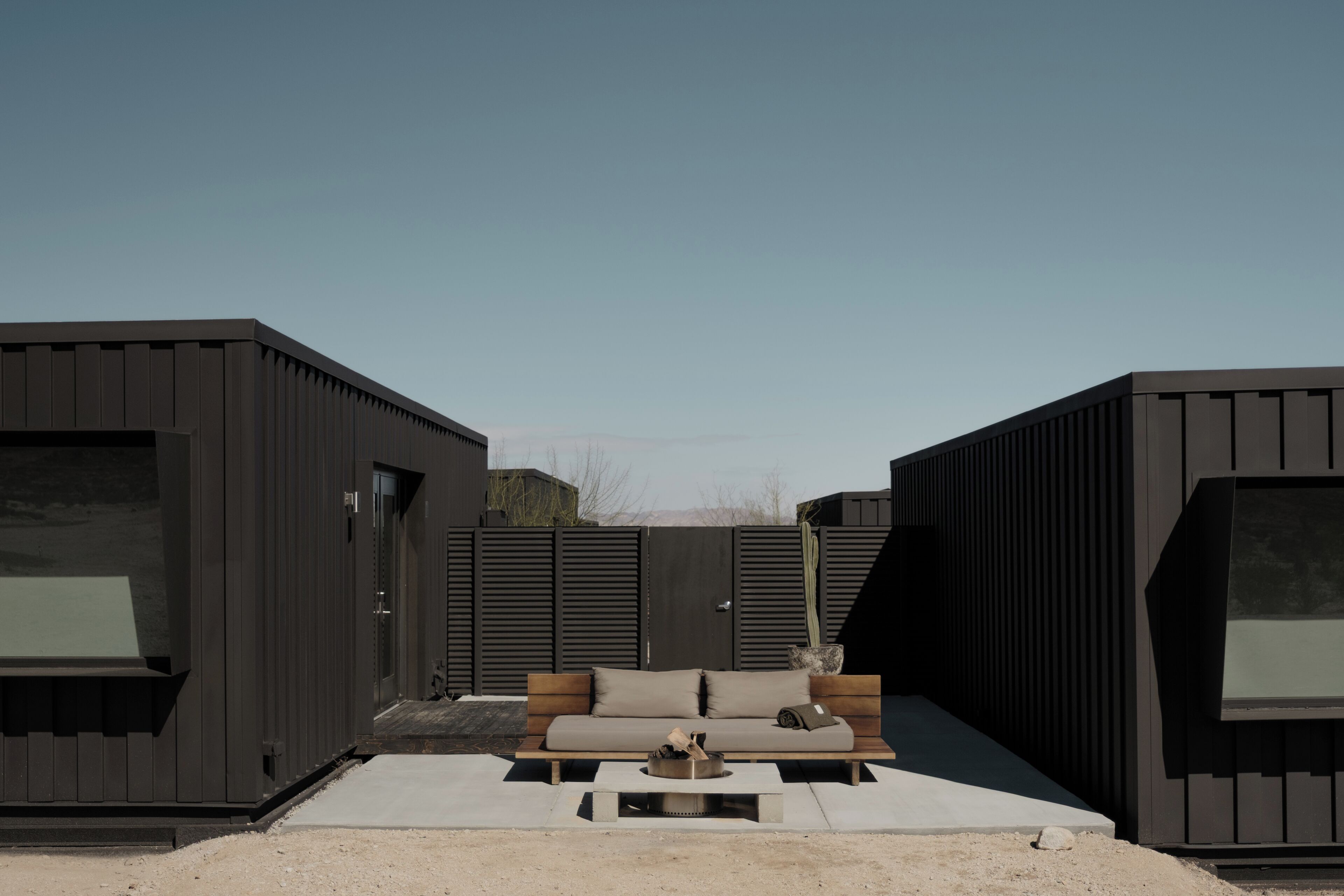 Un espacio exterior moderno con área de estar, que incluye un sofá y una mesa de centro, rodeado de edificios de tonos oscuros.