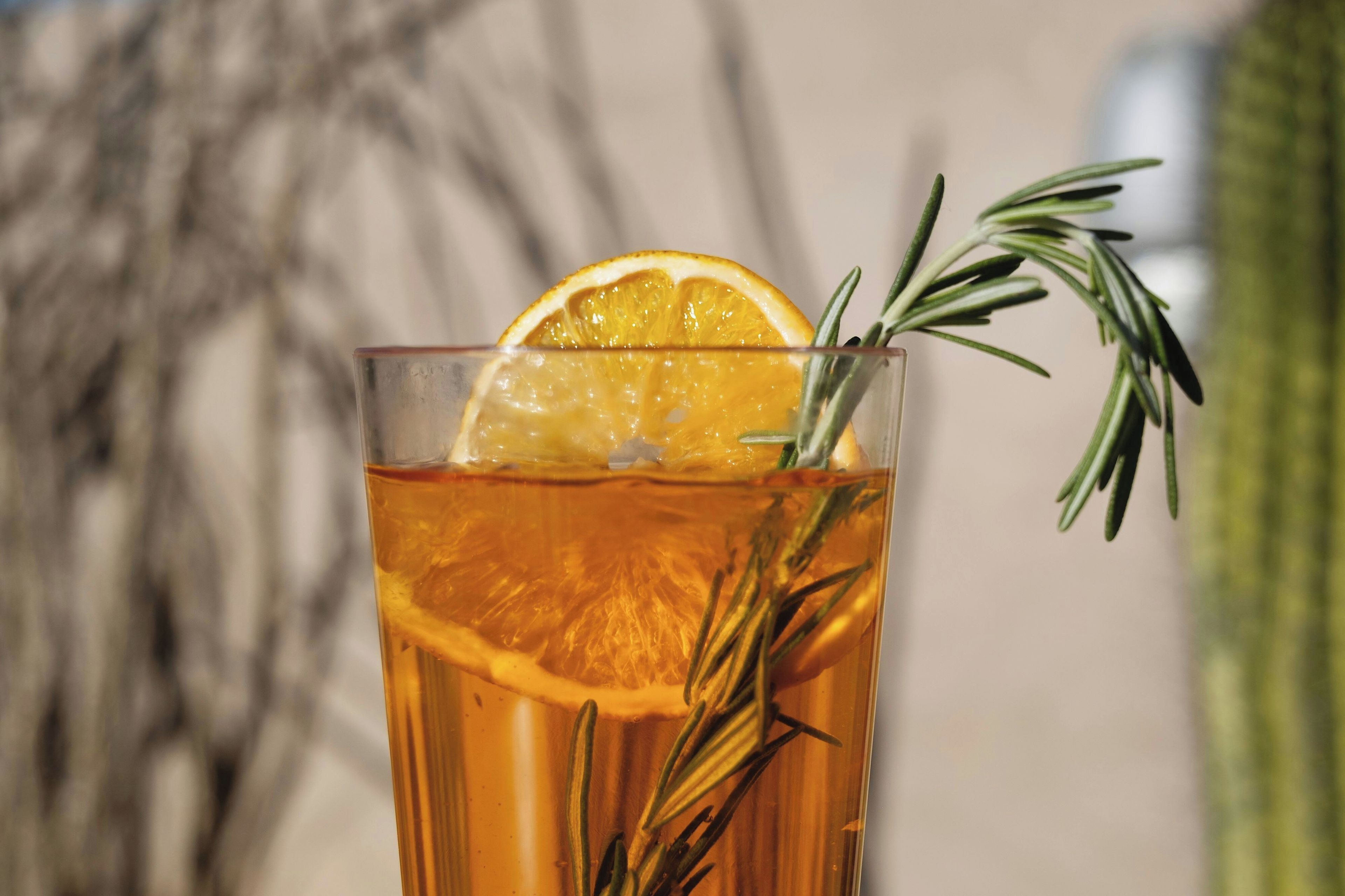 A glass of amber-colored liquid with a slice of orange and a sprig of rosemary.
