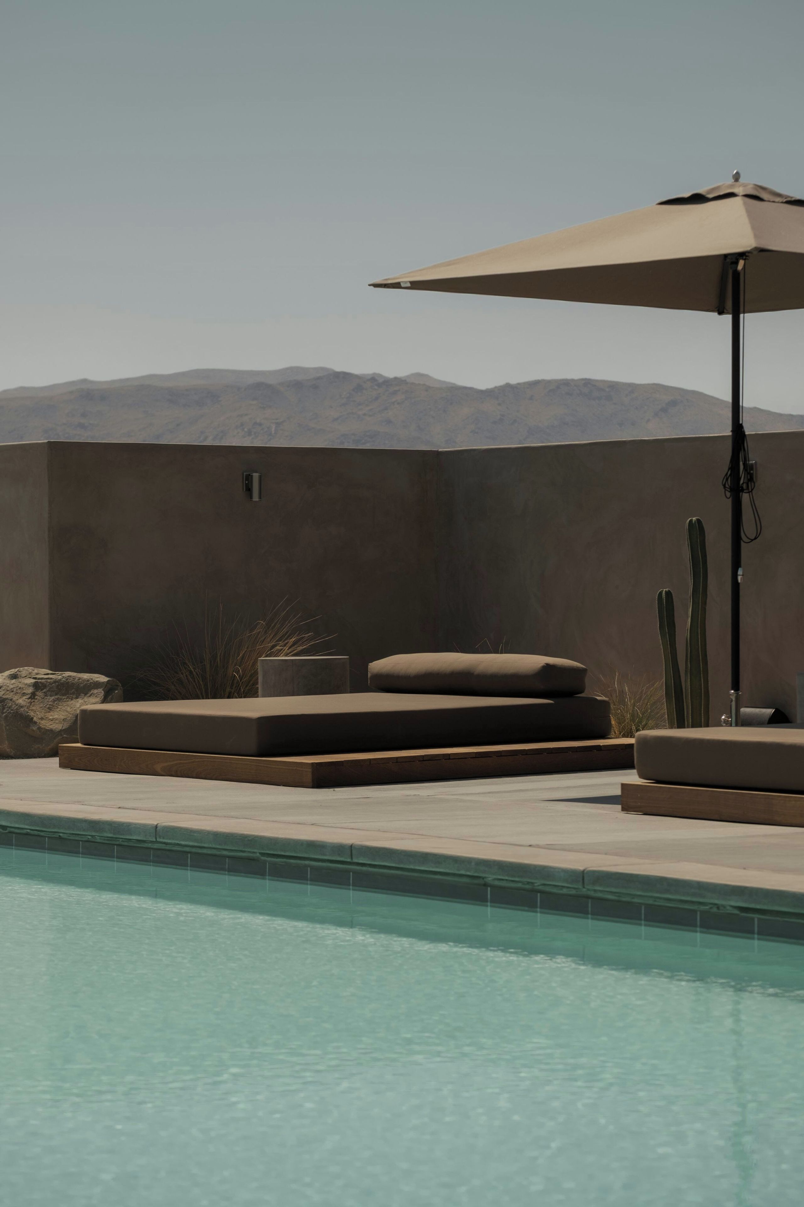 A pool area with a lounge chair and an umbrella, a cactus, and a view of mountains.