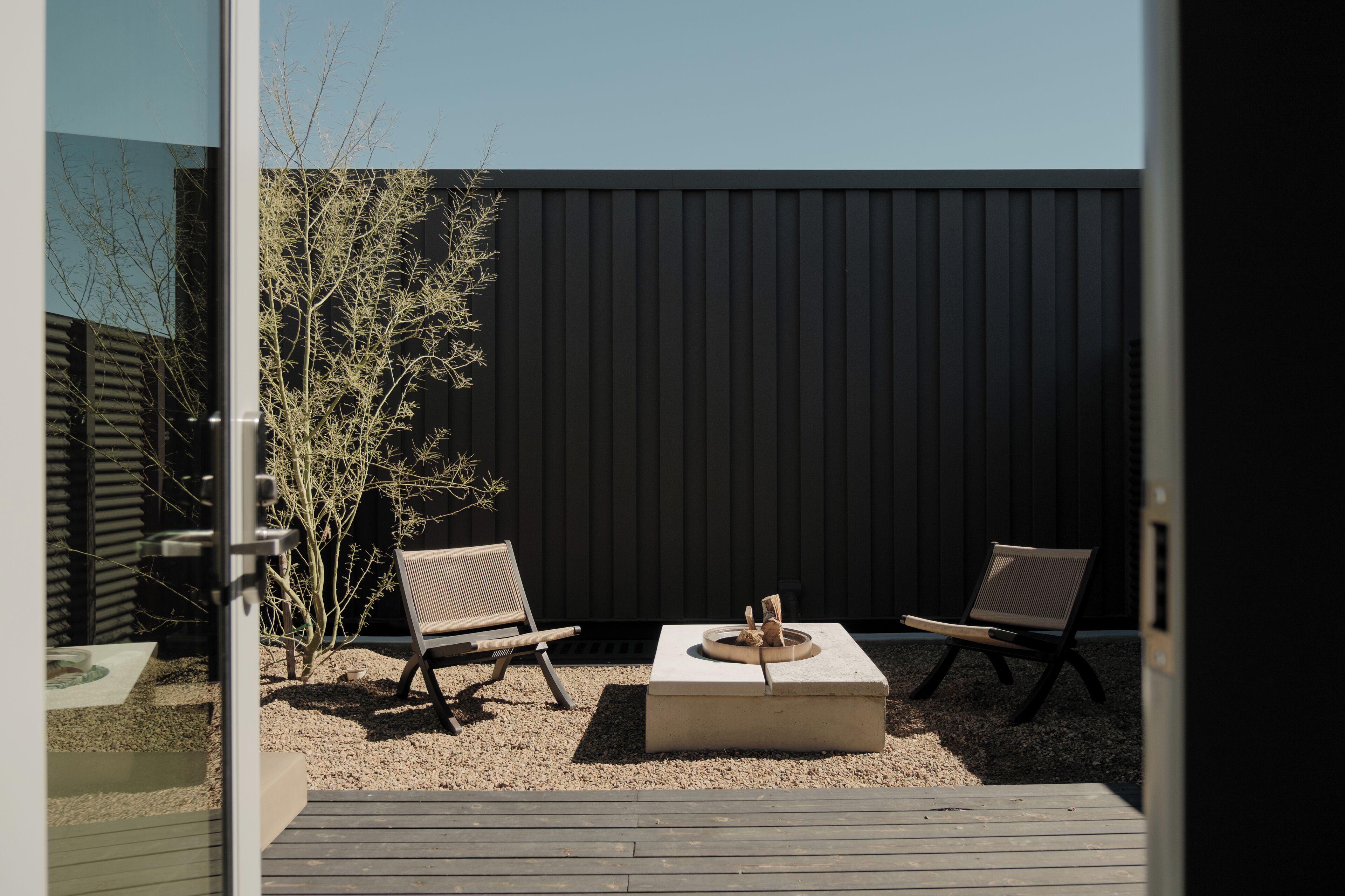 Un espace extérieur aménagé avec deux chaises et une table carrée, entouré de gravier et d’un petit arbre.