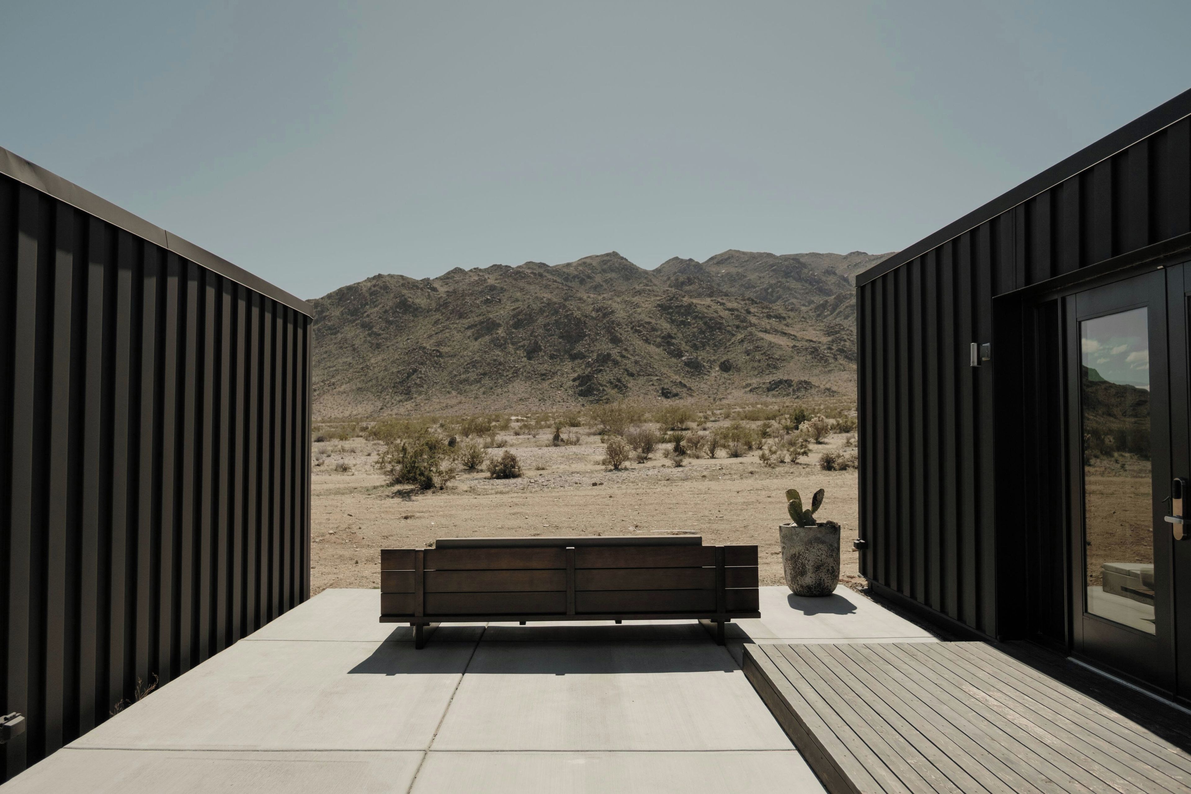 A modern outdoor seating area with a bench and a cactus planter, overlooking a desert landscape with mountains in the distance.