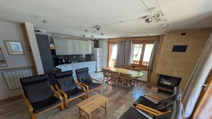 Una sala de estar con un sofá, sillas, una mesa de centro y un comedor con mesa y sillas. Se observa una cocina al fondo con armarios y electrodomésticos.