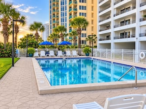 A swimming pool with lounge chairs and umbrellas, surrounded by a white fence, with tall buildings in the background.