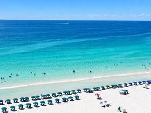 A beach with clear turquoise water, people swimming, and rows of umbrellas on the sand.