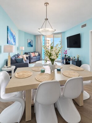 A modern living room with a dining area, featuring a large table with white chairs, a sofa, a TV, and artwork on the walls.