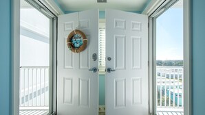 A white door with a 'Welcome' sign and a basket on the wall, leading to a balcony with a view of buildings. - Sunsets on the Sound + Walk to the Beach (Atlantic Beach)