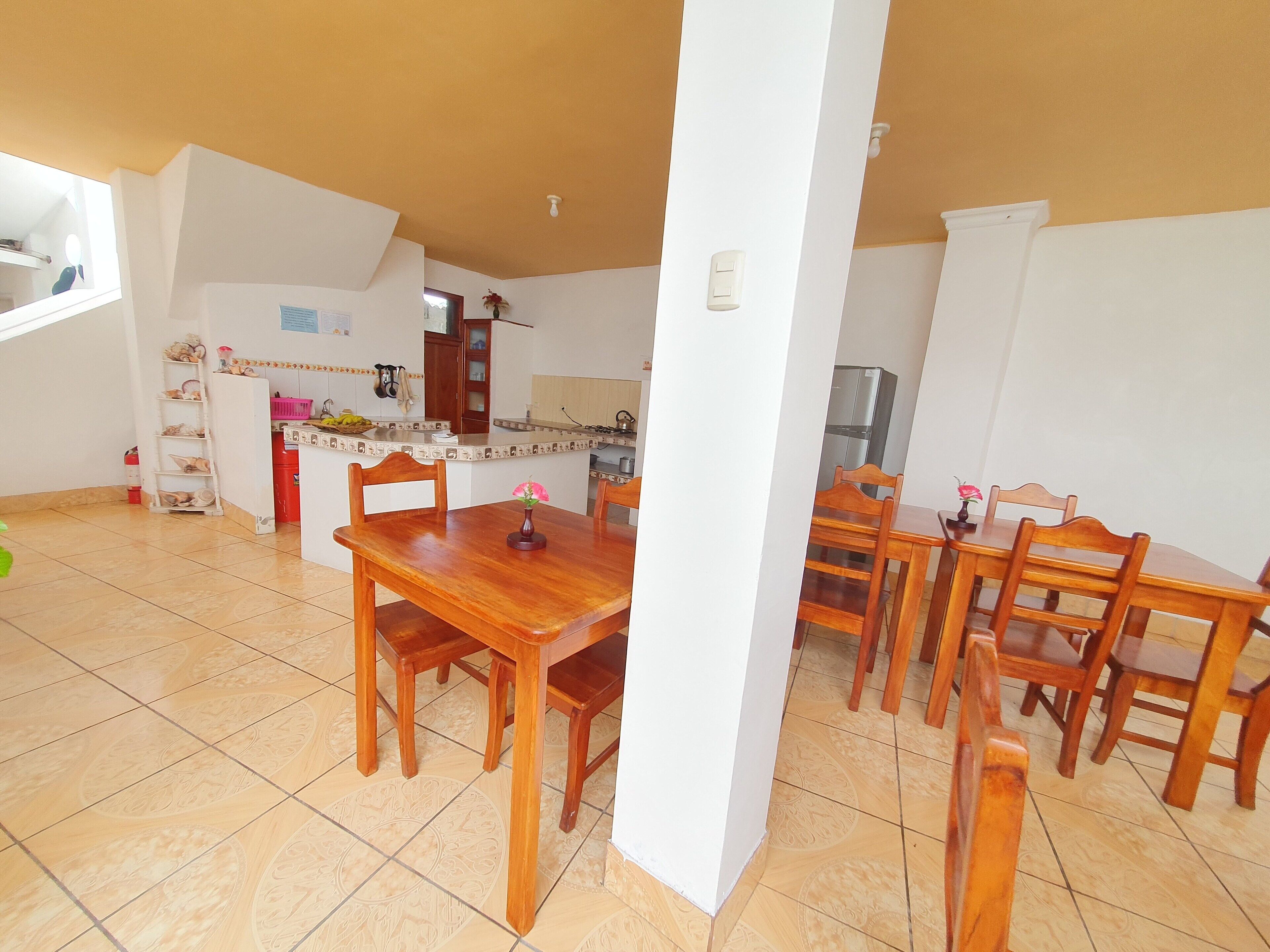 A dining area with a wooden table and chairs, a kitchen in the background, and a white pillar in the foreground.