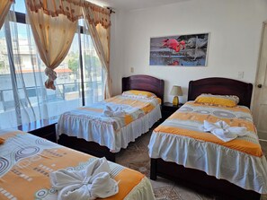 A hotel room with two beds, a window with curtains, a nightstand with a lamp, and a painting of a flamingo. - Cerro Azul (Puerto Villamil)