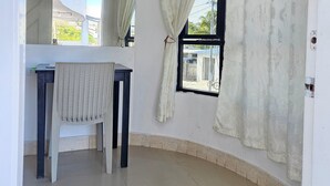 Una habitació amb terra de rajola, una taula de treball amb una cadira, una finestra amb cortines i una làmpada al sostre.
