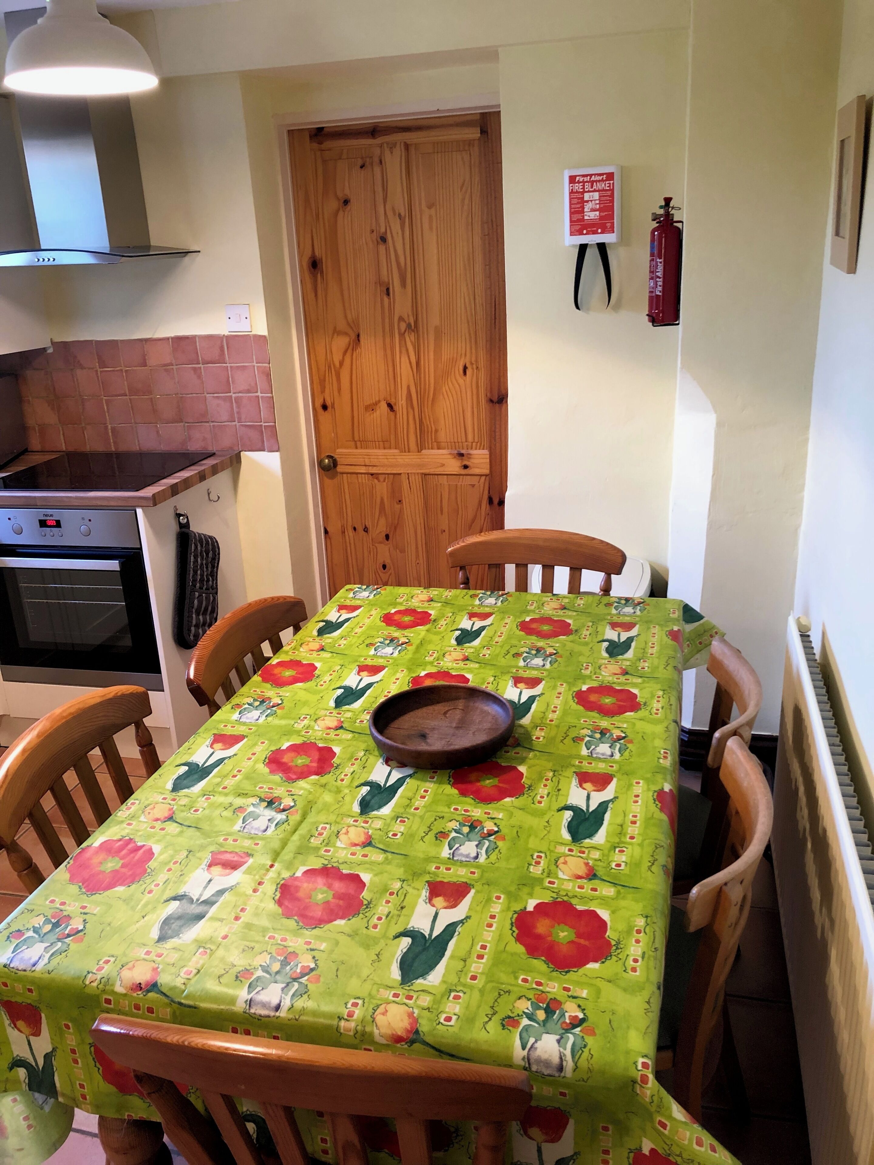 A dining area with a table covered in a floral tablecloth, wooden chairs, a fire extinguisher on the wall, and a kitchen with a stove and oven.