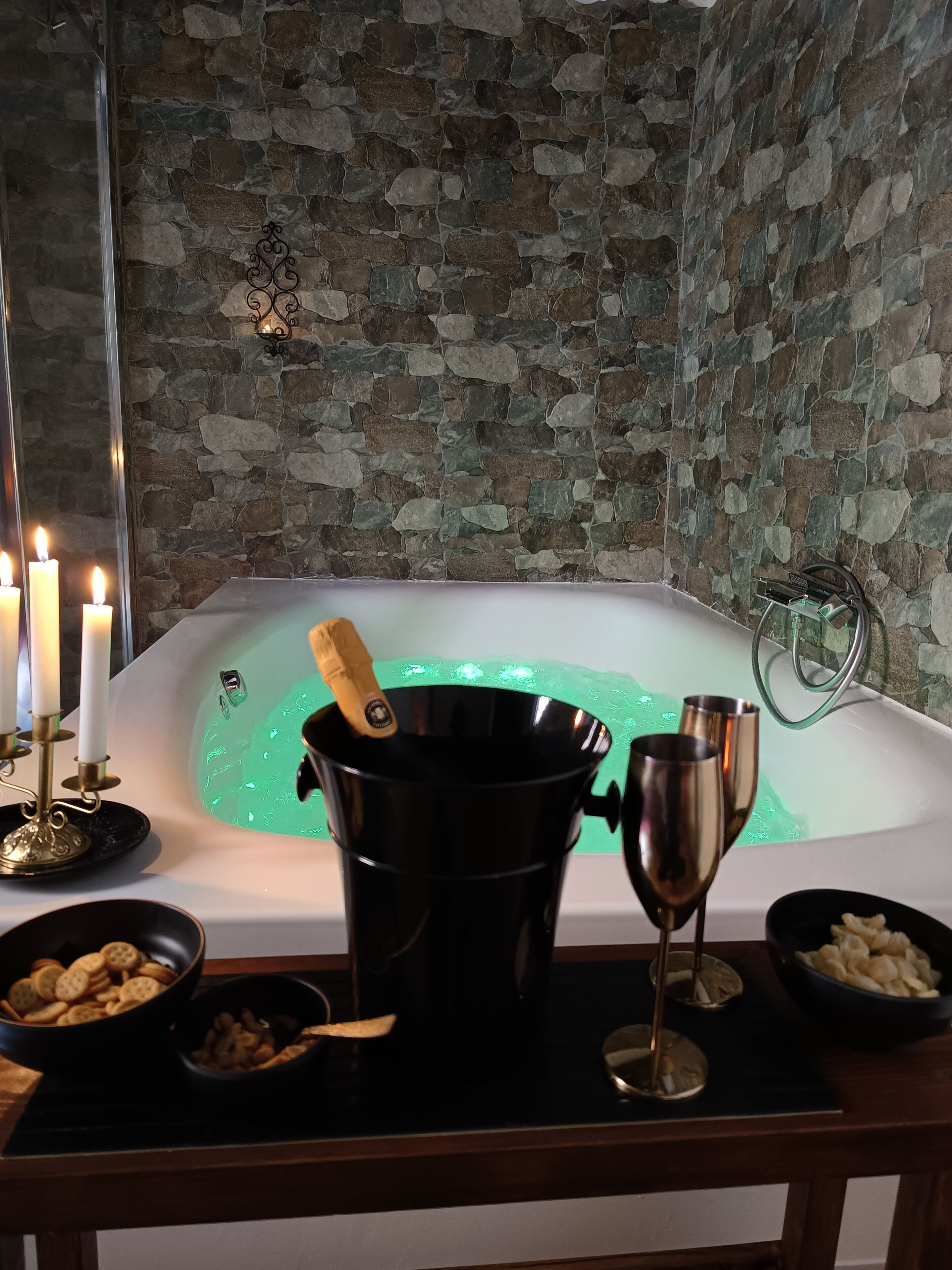 A room with a stone wall, a bathtub with green lighting, candles, a champagne bottle, and snacks.
