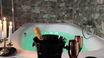 A room with a stone wall, a bathtub with green lighting, candles, a champagne bottle, and snacks.