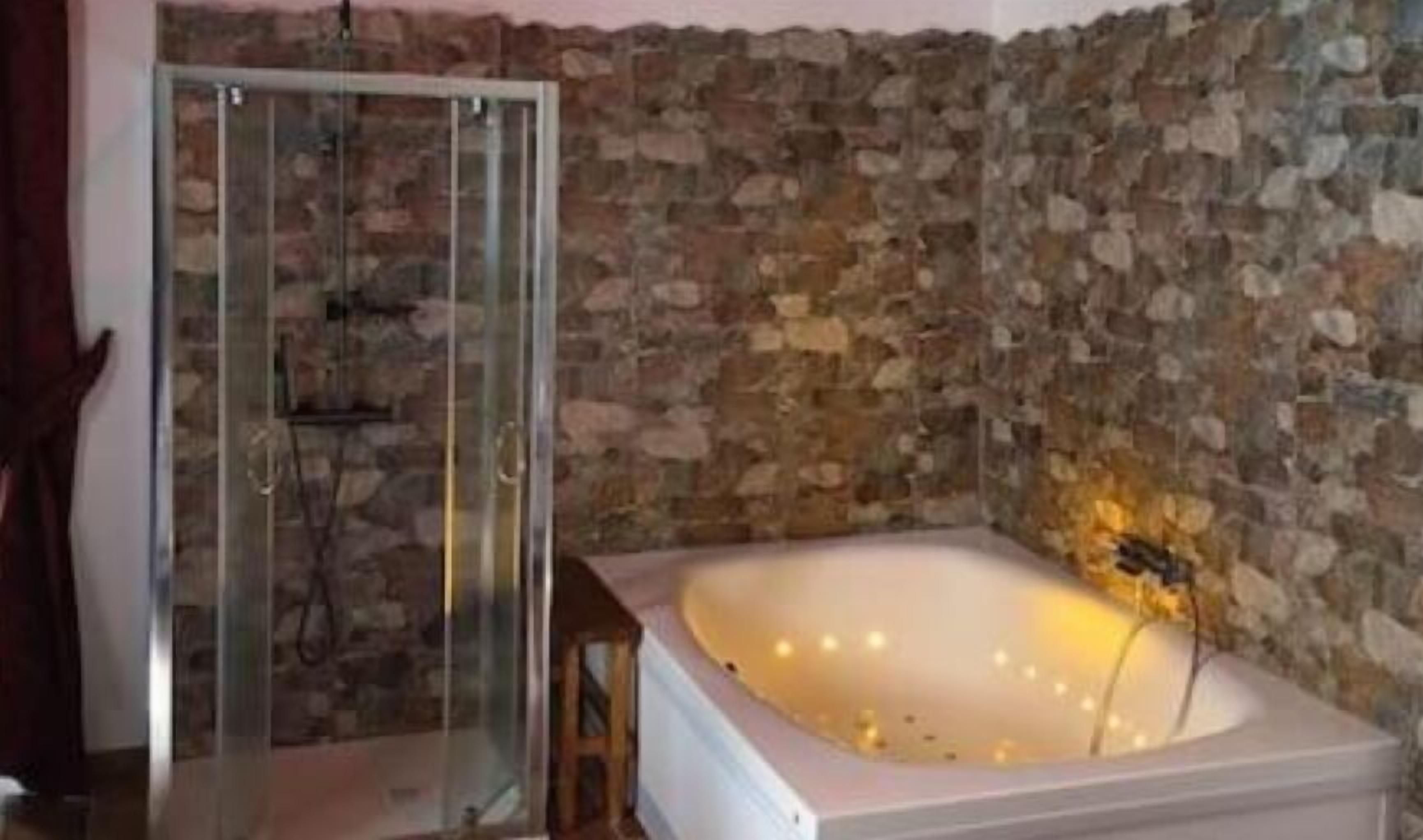 A bathroom with a stone wall, a glass shower enclosure, and a white bathtub with a golden light fixture.