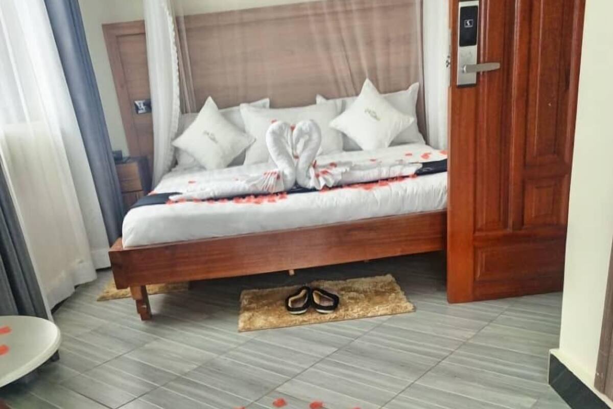 Una cama bien hecha con sábanas blancas y un cabecero de madera, una puerta de madera con manija, y un piso de baldosas con pétalos de rosa esparcidos.