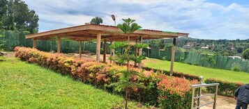 Un espacio al aire libre cubierto, con una estructura de madera, rodeado de setos cuidados y una pendiente cubierta de césped.