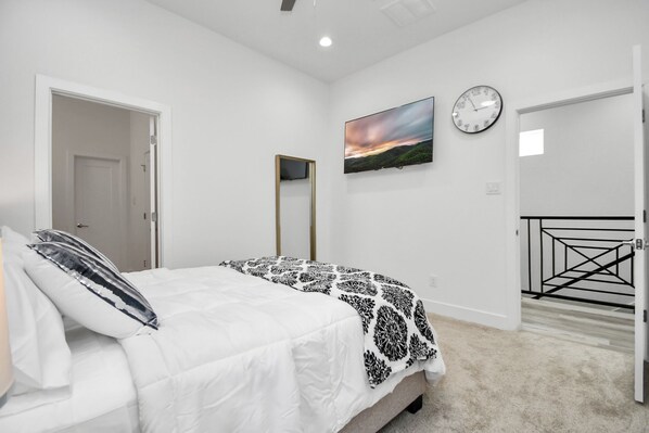 A bedroom with a bed, a wall-mounted TV, a clock, and a door leading to a staircase. - Opal Oasis Lodging - MedCenter - Downtown
(Houston)