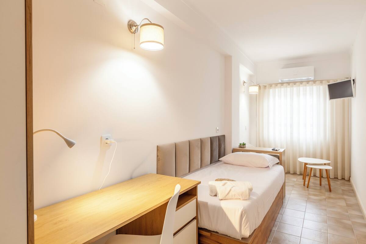 Une chambre d’hôtel comprenant un lit, un bureau, une chaise, une table de chevet, une lampe, un téléviseur fixé au mur et une fenêtre avec des rideaux.
