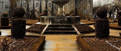 Ein Innenhof mit Brunnen, symmetrisch geschnittenen Hecken und einem historischen Gebäude mit Zinnen im Hintergrund.