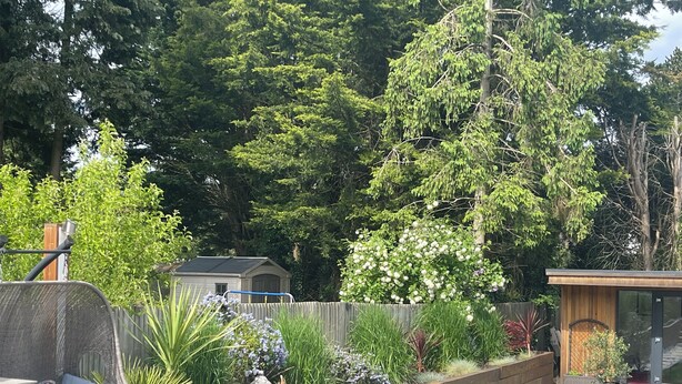 Un jardin à l’arrière de la maison, une terrasse en bois, un petit abri de jardin et un espace aménagé avec une chaise et une table.