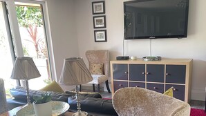 Un salon avec un téléviseur fixé au mur, une table basse en bois avec une lampe, un fauteuil beige et un canapé de couleur foncée.