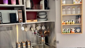 Une cuisine compacte équipée d’un évier en acier inoxydable, d’un four à micro-ondes et d’une bouilloire. On y trouve des étagères remplies de divers ustensiles de cuisine et un petit meuble en bois contenant des bocaux.
