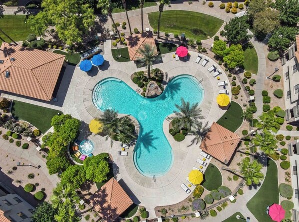 An aerial view of a resort with a central swimming pool, surrounded by palm trees, lounge chairs, and umbrellas.