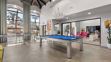 Una mesa de billar en una habitación moderna con ventanas amplias, una zona de bar y un espacio de estar con sillas rojas.