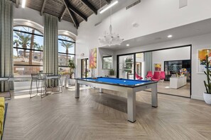 A pool table in a modern room with large windows, a bar area, and a seating space with red chairs.