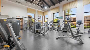 Un gimnasio con diversas máquinas de ejercicio, ventanas amplias y un techo con una lámpara de diseño geométrico.