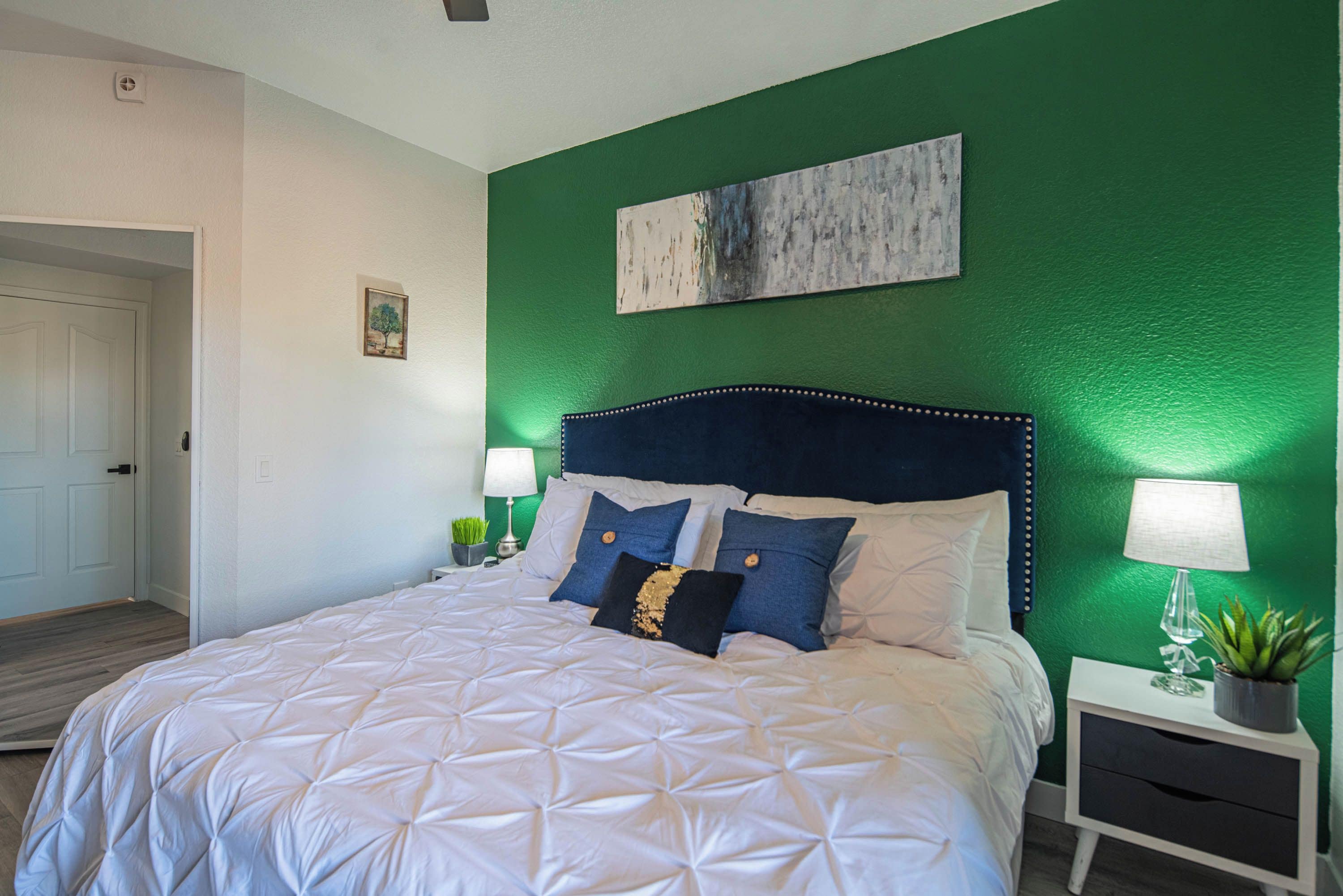 Una cama bien hecha con colcha blanca, dos almohadas y un cabecero con diseño decorativo. Hay una mesita de noche con una lámpara y una planta pequeña.
