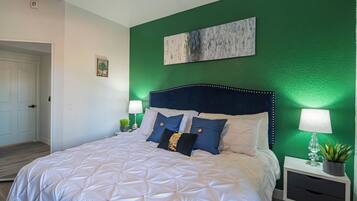 Una cama bien hecha con colcha blanca, dos almohadas y un cabecero con diseño decorativo. Hay una mesita de noche con una lámpara y una planta pequeña.