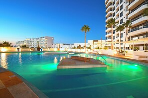 A swimming pool with a wooden deck and lounge chairs, surrounded by palm trees and modern buildings.